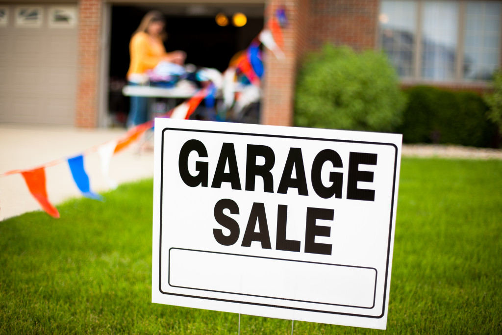 Garage sale sign on the front yard of a suburban house with a woman looking at items on a table.